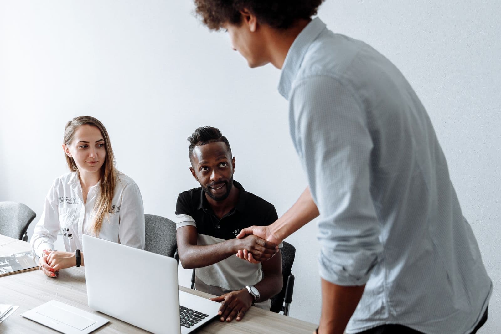 Two colleagues shaking hands in an office, with two more people seated at a laptop nearby